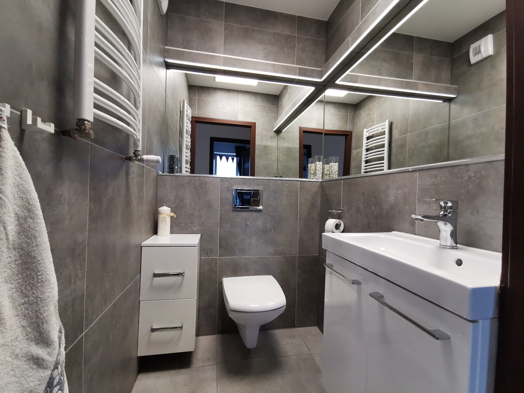 Nowoczesna łazienka wykończona w szarościach z białymi meblami, lustrem z podświetleniem LED i grzejnikiem drabinkowym, widok na otwarte drzwi.