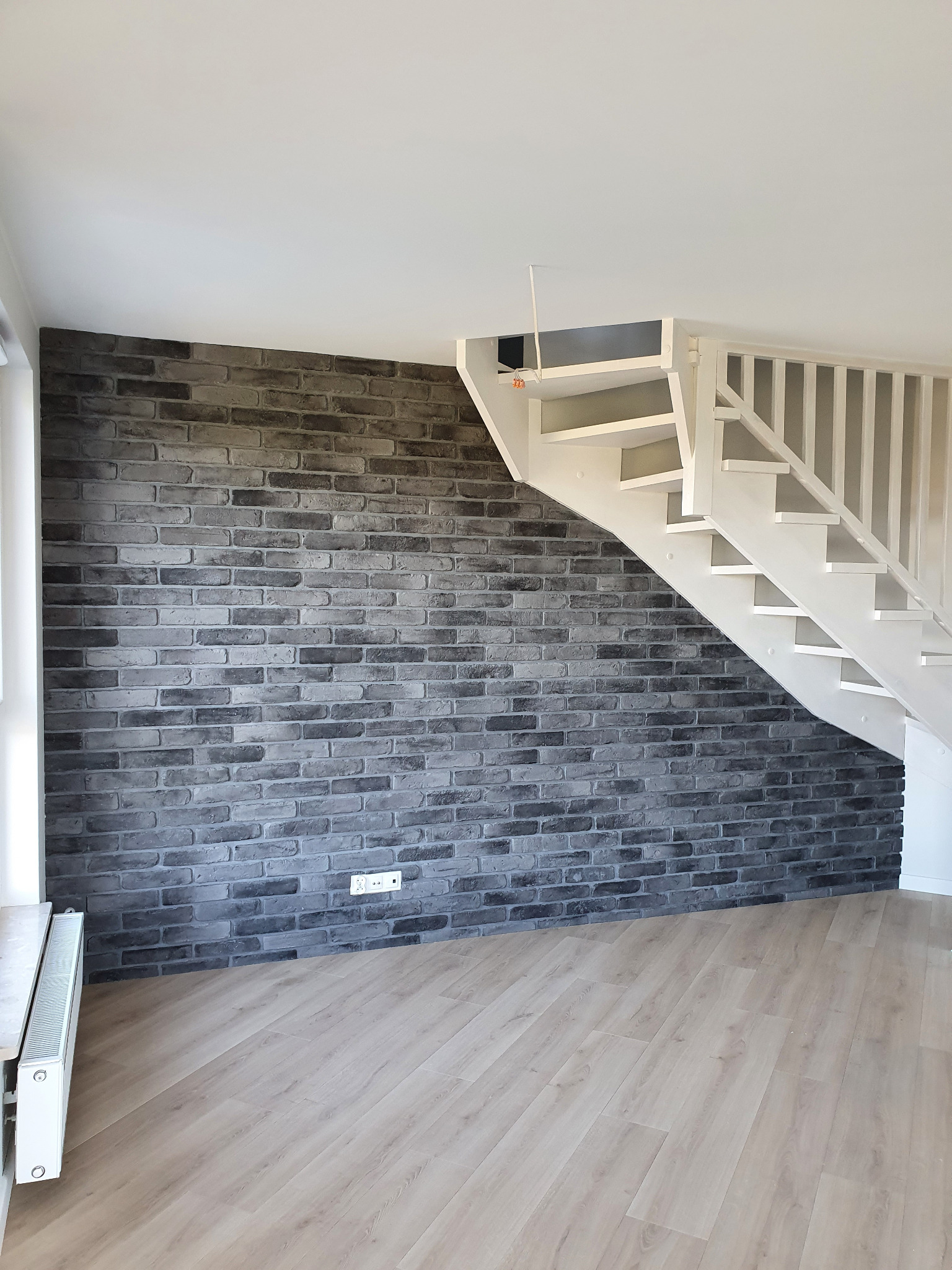 Wykończone wnętrze mieszkania: ściana z szarej cegły dekoracyjnej, białe drewniane schody z barierką i jasna podłoga z paneli imitujących drewno, widoczny grzejnik.