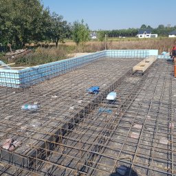 PAWEŁ SIENKIEWICZ SIENKIEWICZ CONSTRUCTON - Siatka zbrojeniowa fundamentów z termoizolacją obwodową, widoczni pracownicy w tle, butelka wody i cegły na siatce.