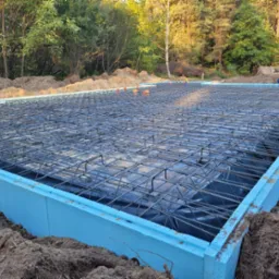 Zbrojenie płyty fundamentowej w szalunku z niebieskiego styropianu, widok z góry na siatkę z prętów stalowych ułożoną na czarnej folii, w tle zalesiony teren.