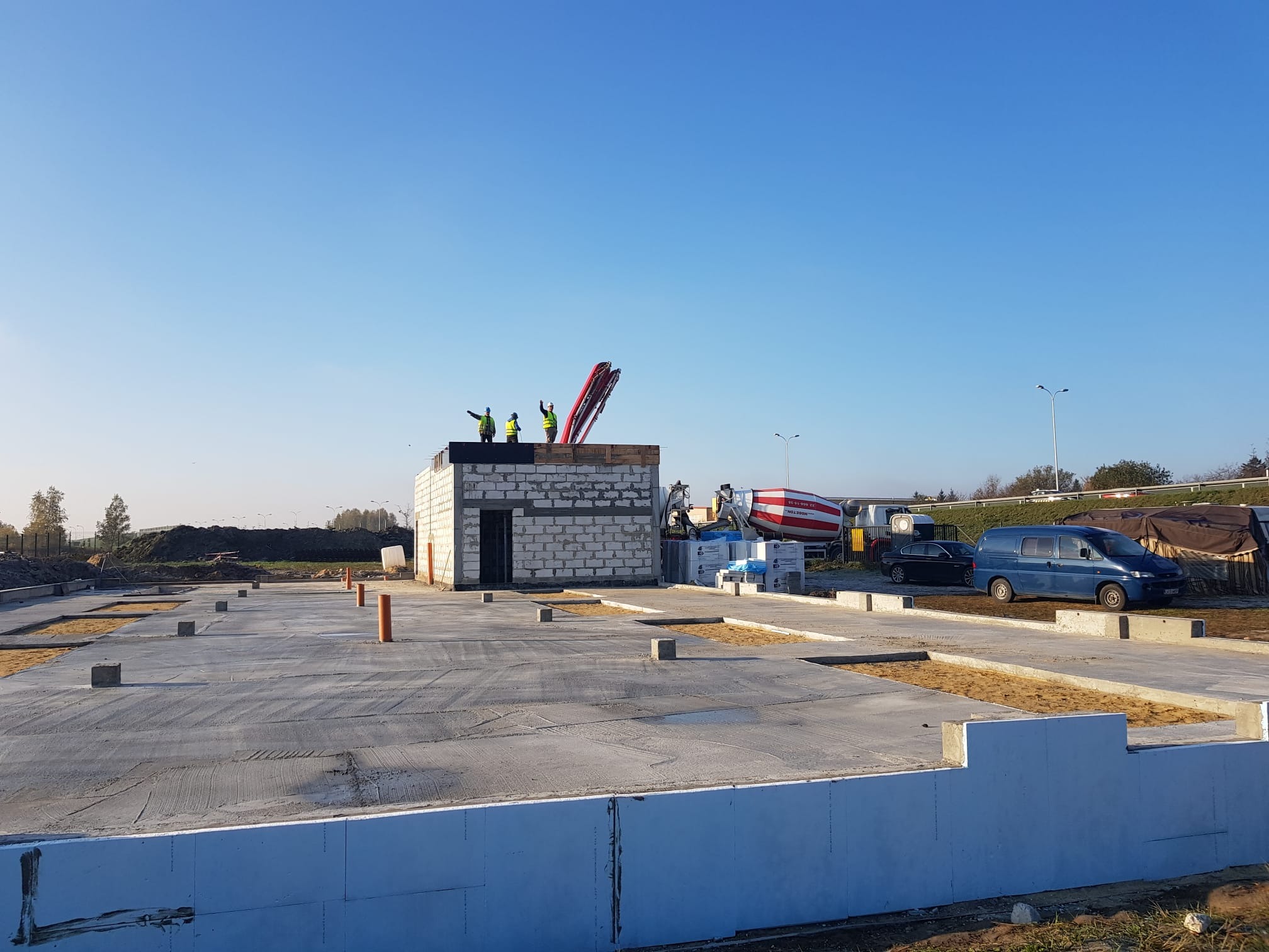 Wylana płyta fundamentowa z widocznymi izolacjami pionowymi, blokami betonowymi, rurami instalacyjnymi i ekipą pracowników na dachu budynku w trakcie budowy, przy słonecznej pogodzie.