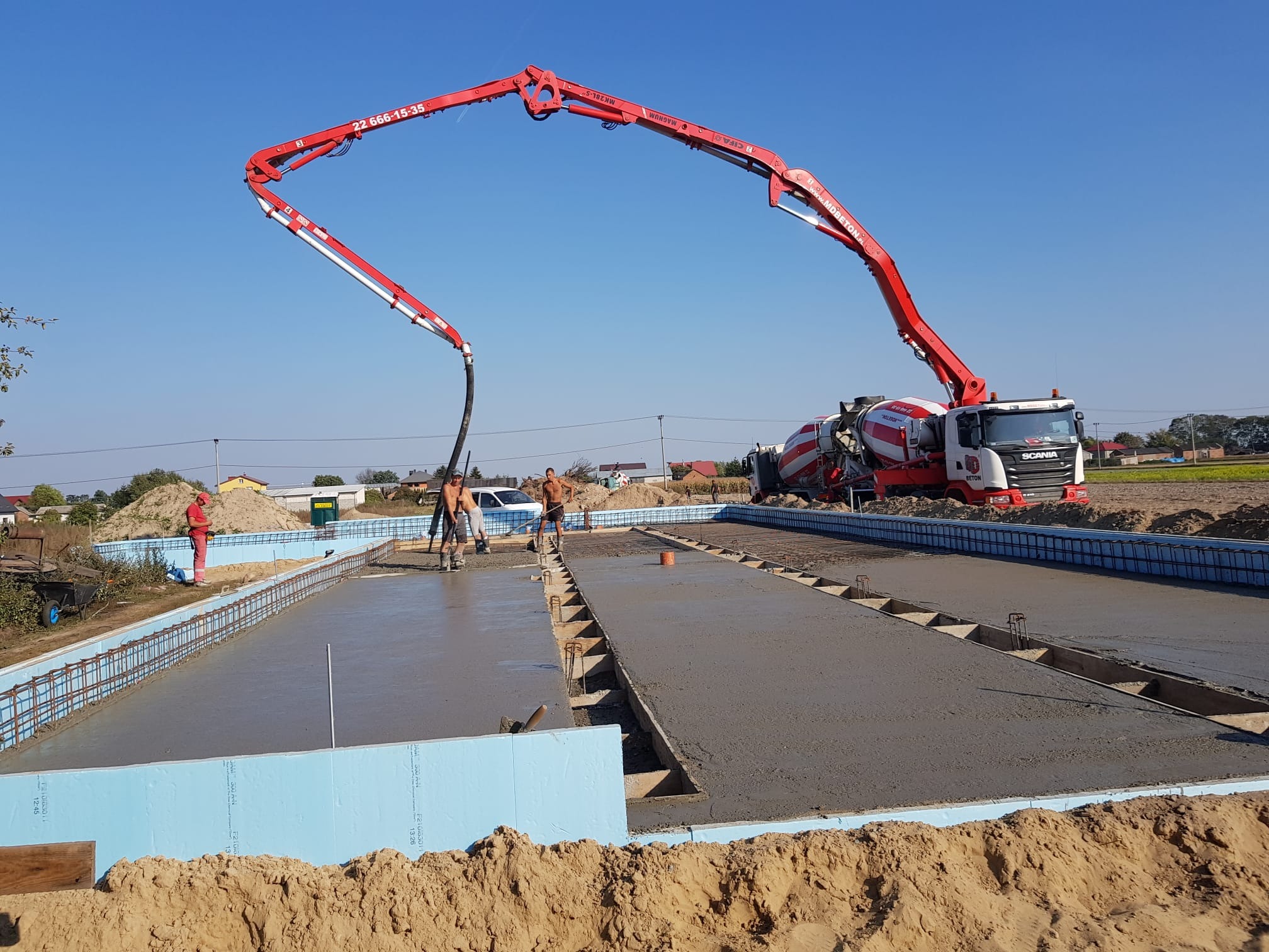 Wylewanie betonu na przygotowany zbrojony fundament z użyciem pompy do betonu, widoczny mikser do betonu Scania i pracownicy w trakcie pracy.