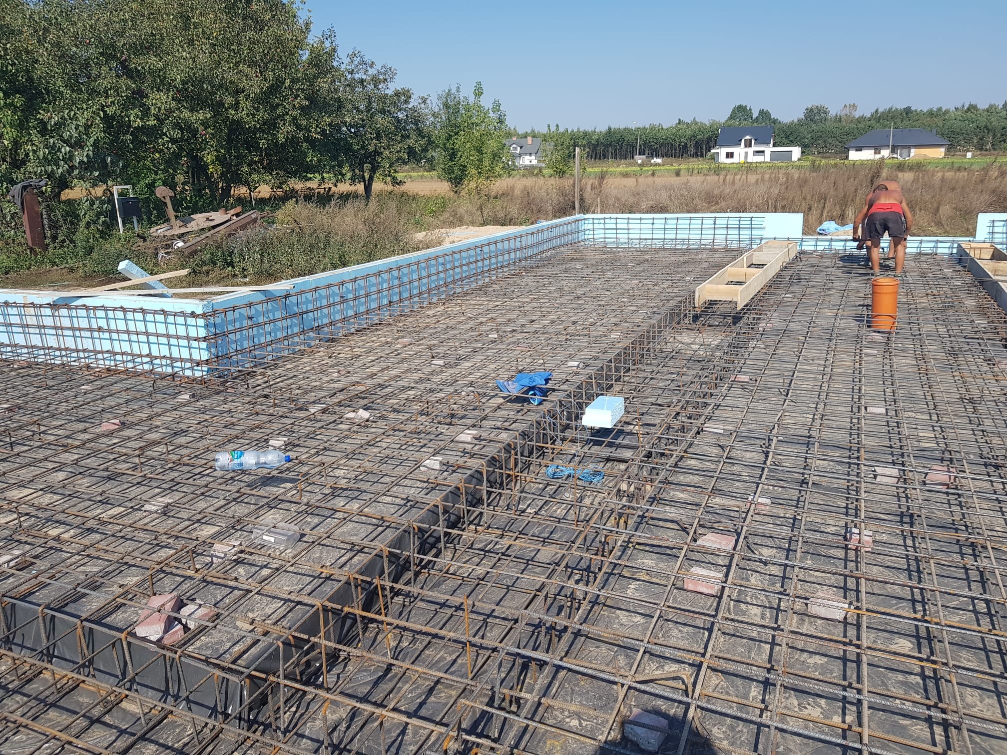 Siatka zbrojeniowa fundamentów z termoizolacją obwodową, widoczni pracownicy w tle, butelka wody i cegły na siatce.