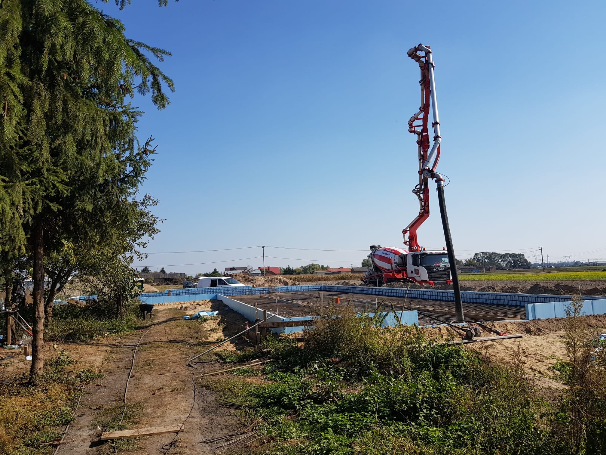Wylewanie betonu do fundamentów pod budynek za pomocą pompy do betonu Scania, widoczne niebieskie szalunki i zbrojenie.
