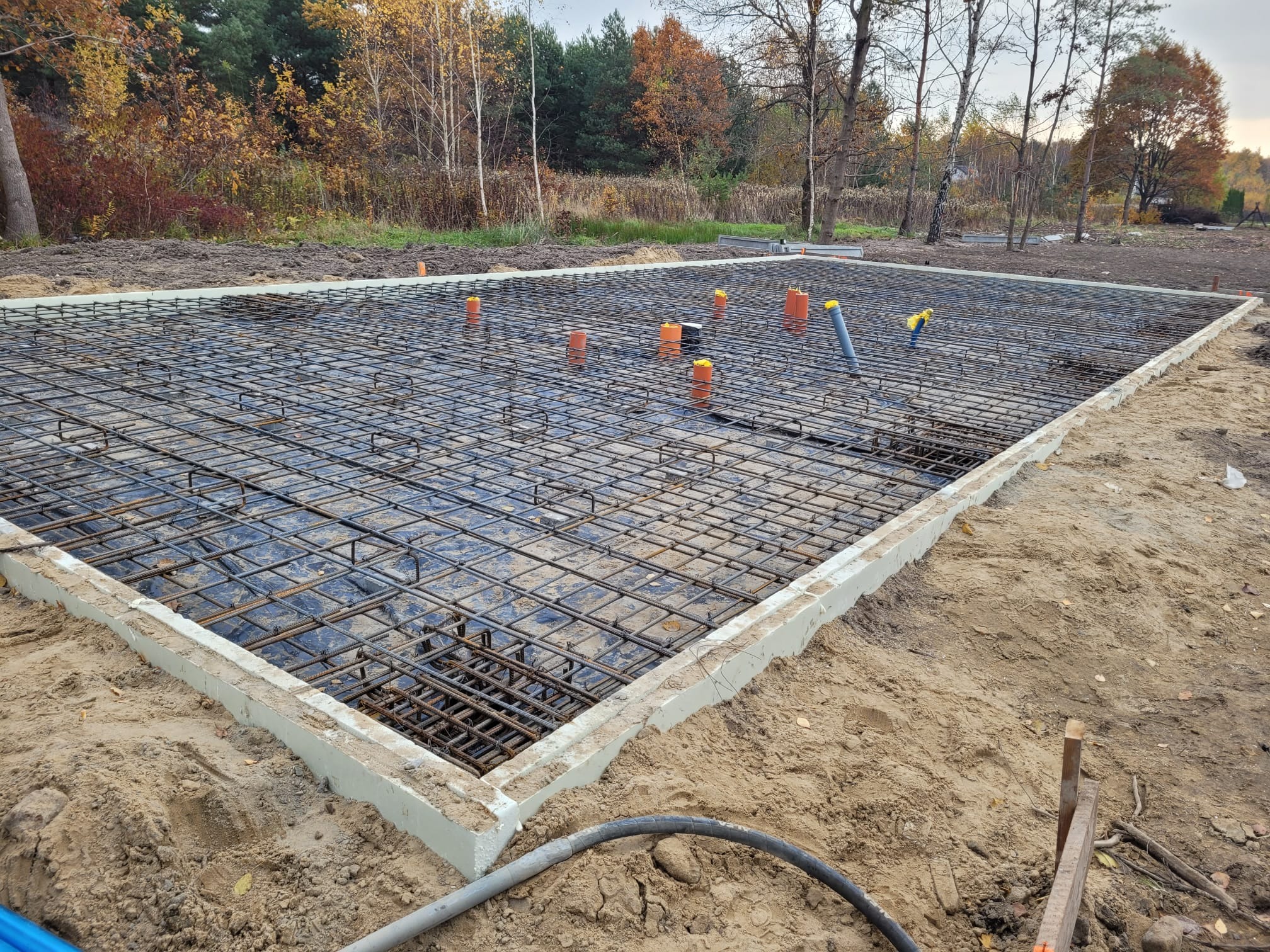 Przygotowany szalunek z siatką zbrojeniową na płycie fundamentowej z wystającymi rurami instalacyjnymi w kolorze pomarańczowym i szarym, na piaszczystym terenie budowy z widocznymi drzewami w tle.