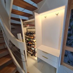 GKM MEBLE Sławomir Maciejczyk - Biała szafa wnękowa z półkami na buty i wieszakami pod schodami spiralnymi z drewnianymi stopniami, oświetlona listwą LED, z dwoma szufladami z złotymi uchwytami.