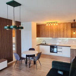 GKM MEBLE Sławomir Maciejczyk - Nowoczesna kuchnia z drewnianymi frontami szafek górnych i białymi dolnymi, okrągły stół z szarymi krzesłami, zielona lampa wisząca i drewniane lamele na ścianie w salonie.