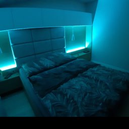 GKM MEBLE Sławomir Maciejczyk - Sypialnia z łóżkiem i tapicerowanym zagłówkiem z podświetleniem LED. Nad łóżkiem szafki wiszące, po bokach małe szafki nocne z lampkami. Całość oświetlona niebieskim światłem.