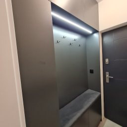 GKM MEBLE Sławomir Maciejczyk - Szara zabudowa w przedpokoju z wbudowanym siedziskiem tapicerowanym, podświetleniem LED i haczykami na ubrania, widok na drzwi wejściowe.