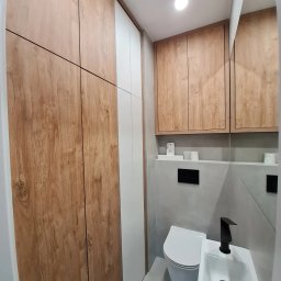 GKM MEBLE Sławomir Maciejczyk - Nowoczesna łazienka z zabudową meblową z płyty laminowanej w kolorze drewna i bieli, szafka nad geberitem, biała umywalka z czarną baterią, szara ściana i lustro.
