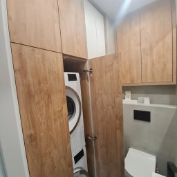 GKM MEBLE Sławomir Maciejczyk - Pralka i suszarka w zabudowie meblowej z drewnopodobnymi frontami, widoczna toaleta i umywalka w małej łazience.