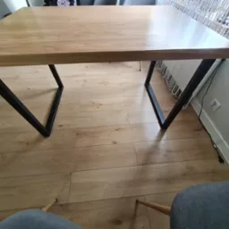 Dębowy stół z czarnymi, metalowymi nogami w kształcie litery V, lakierowany na matowo, z dodatkowymi dostawkami. Widok z góry, parkiet na podłodze.