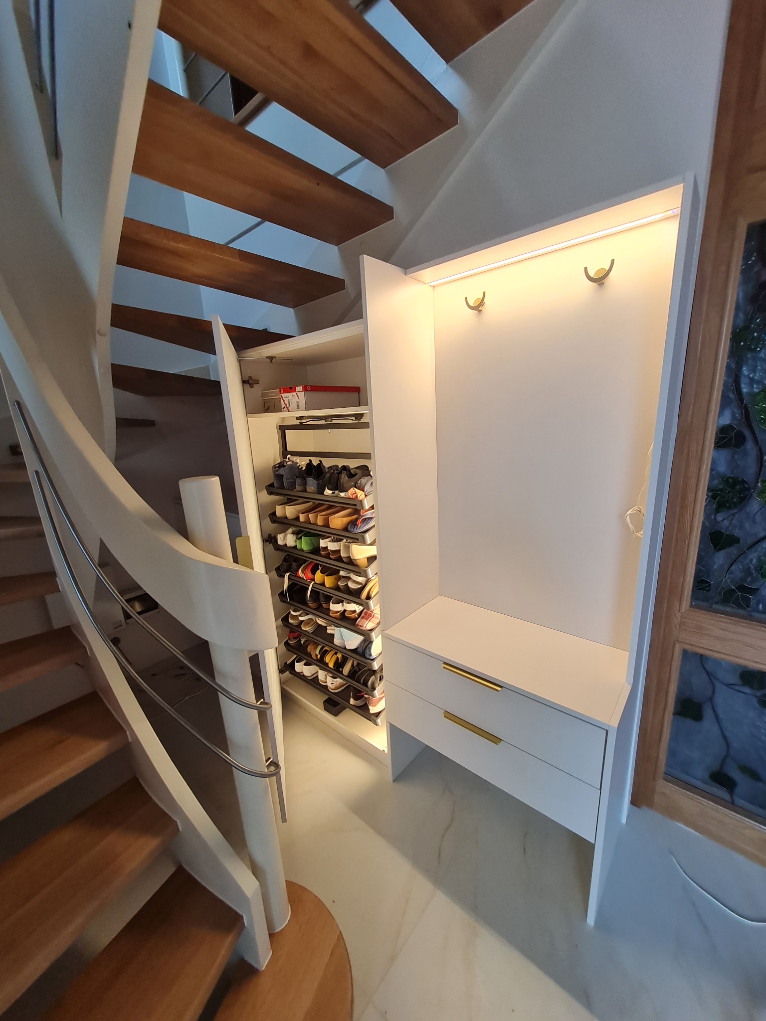 Biała szafa wnękowa z półkami na buty i wieszakami pod schodami spiralnymi z drewnianymi stopniami, oświetlona listwą LED, z dwoma szufladami z złotymi uchwytami.