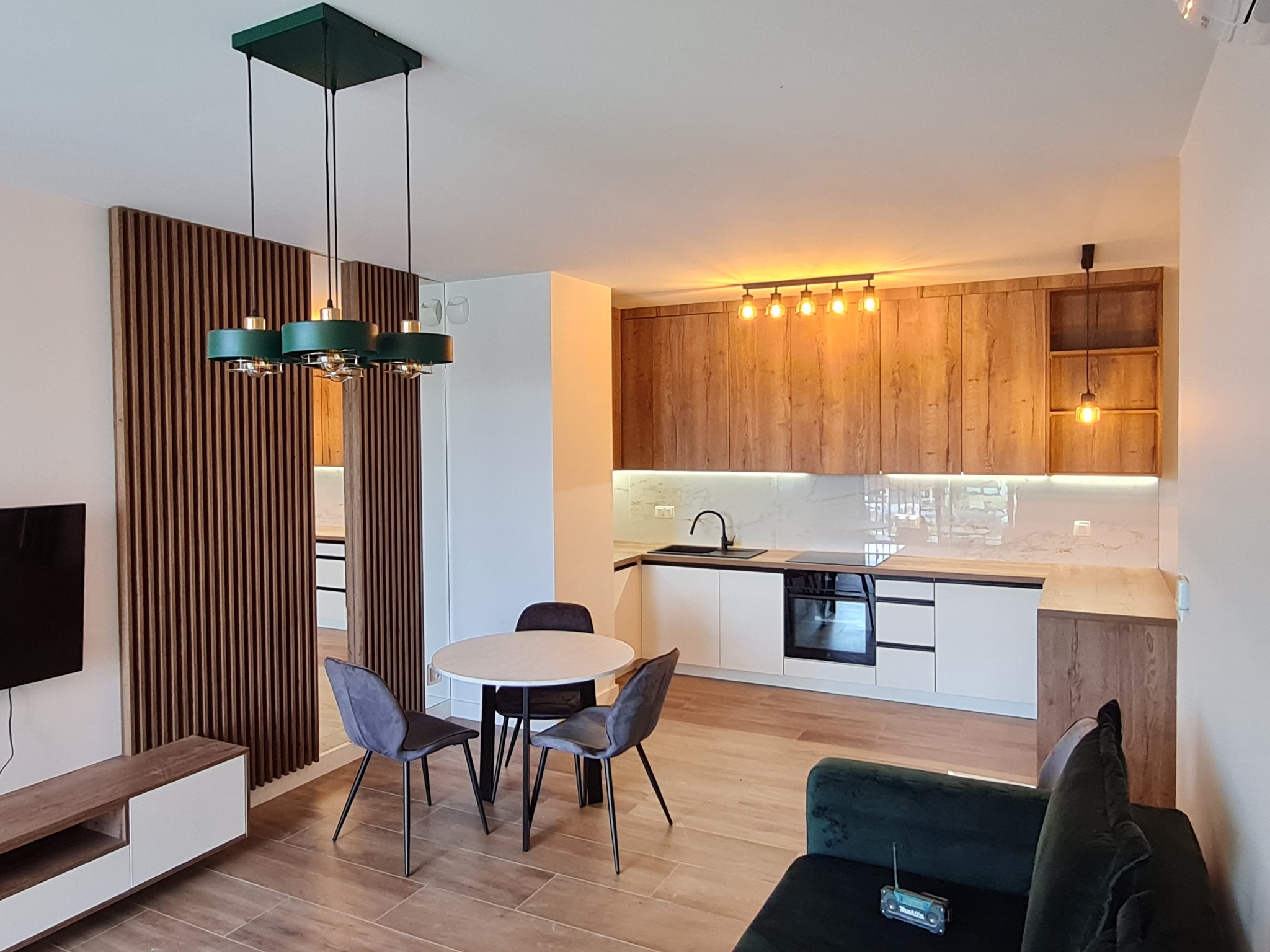 Nowoczesna kuchnia z drewnianymi frontami szafek górnych i białymi dolnymi, okrągły stół z szarymi krzesłami, zielona lampa wisząca i drewniane lamele na ścianie w salonie.