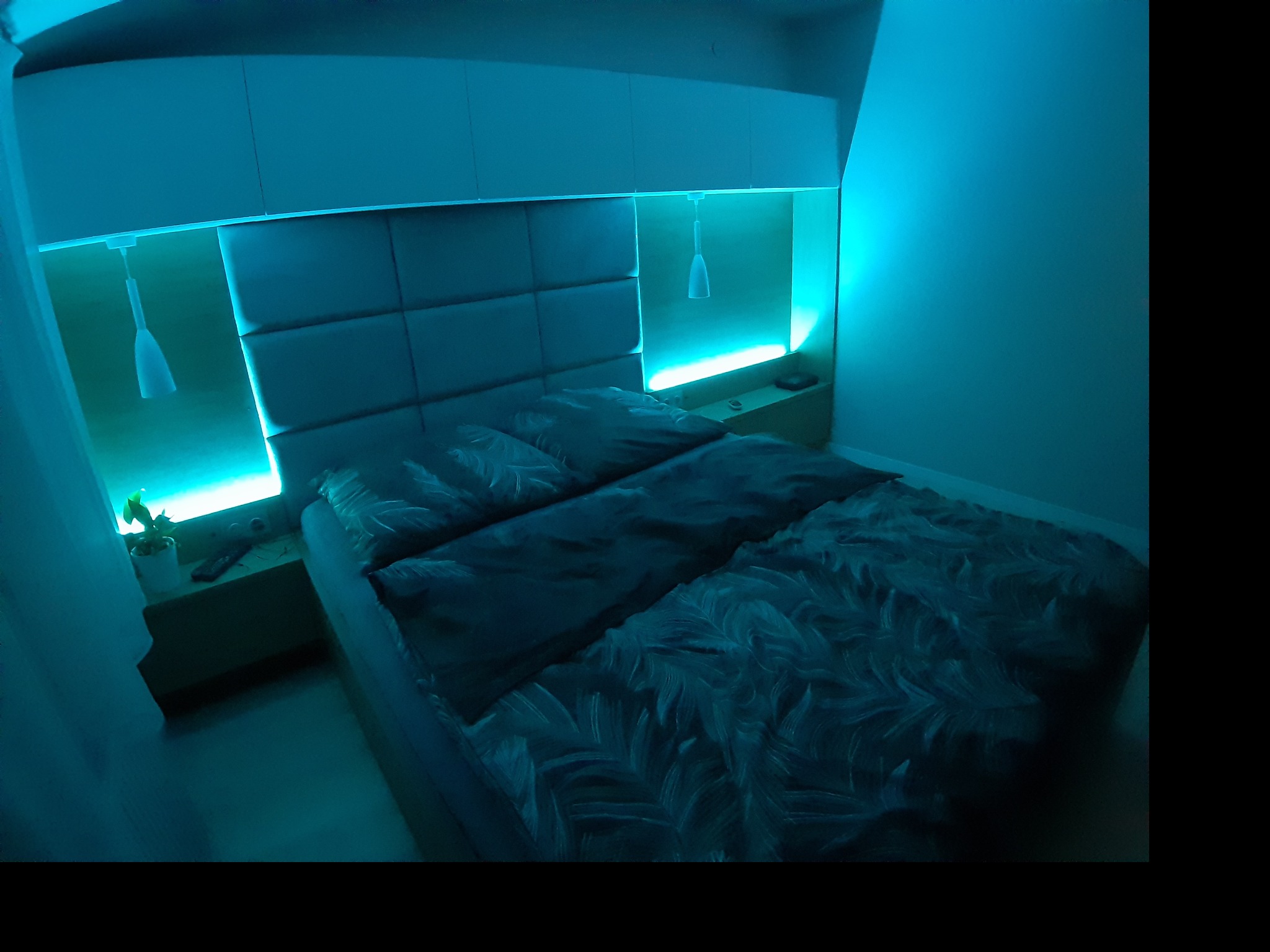 Sypialnia z łóżkiem i tapicerowanym zagłówkiem z podświetleniem LED. Nad łóżkiem szafki wiszące, po bokach małe szafki nocne z lampkami. Całość oświetlona niebieskim światłem.
