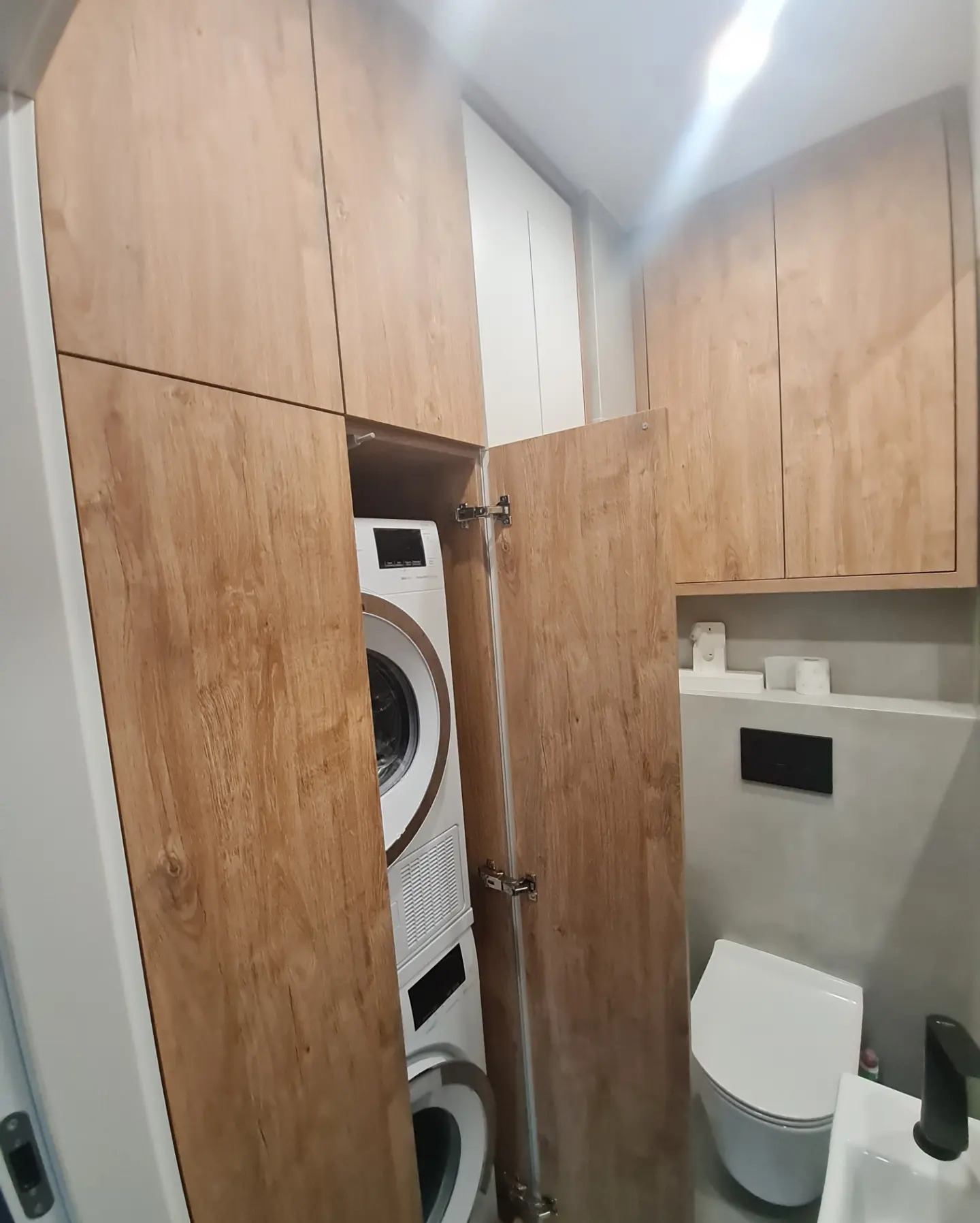 Pralka i suszarka w zabudowie meblowej z drewnopodobnymi frontami, widoczna toaleta i umywalka w małej łazience.