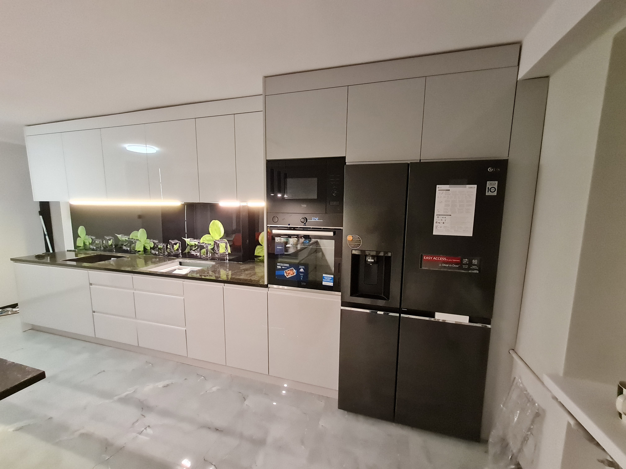 Nowoczesna kuchnia z białymi, błyszczącymi frontami szafek, czarnym blatem i szklanym panelem nad blatem z motywem roślinnym, z zabudowaną czarną lodówką i piekarnikiem.