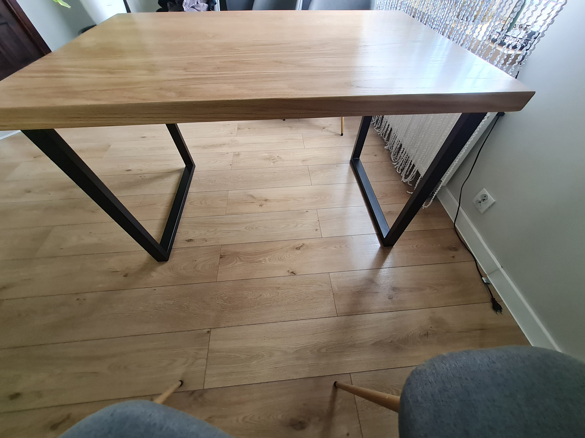Dębowy stół z czarnymi, metalowymi nogami w kształcie litery V, lakierowany na matowo, z dodatkowymi dostawkami. Widok z góry, parkiet na podłodze.