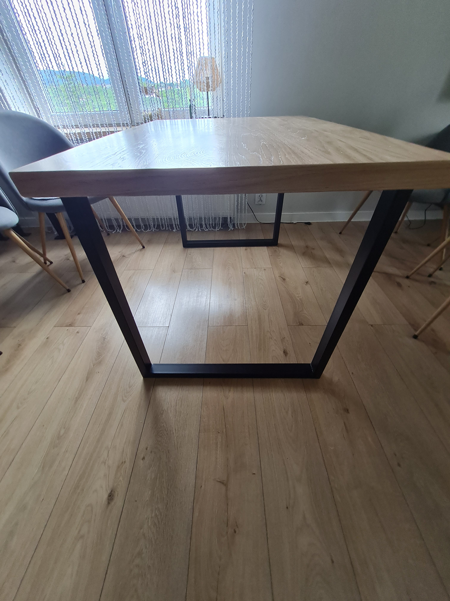Stół dębowy z blatem lakierowanym na matowo i czarnymi, metalowymi nogami w kształcie trapezu. W tle okno z białymi zasłonami i widok na zieleń.