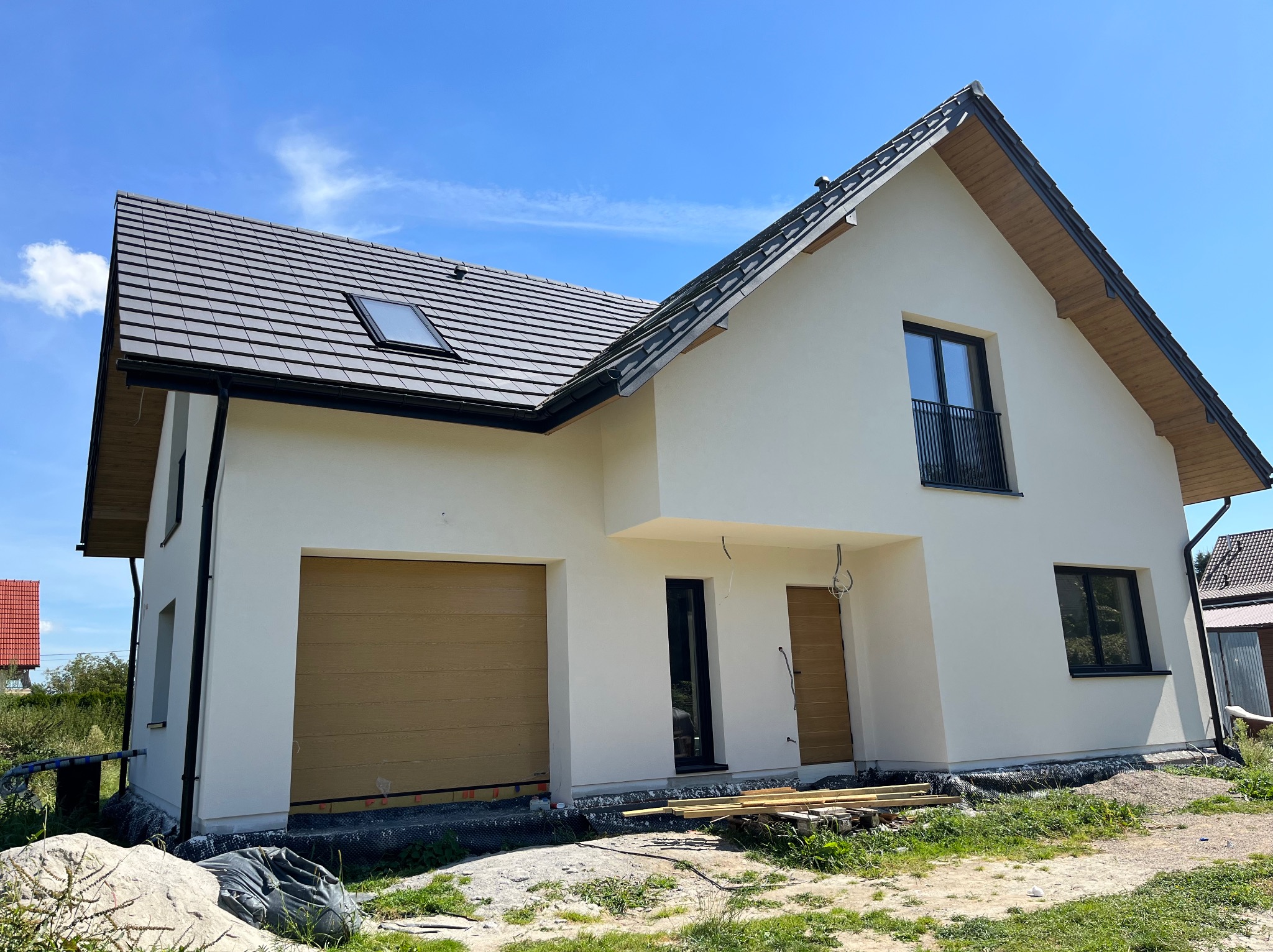 Nowoczesny dom jednorodzinny z jasną elewacją i brązową bramą garażową. Widoczne okna z czarnymi ramami, dachówka i fragment niezagospodarowanego terenu.