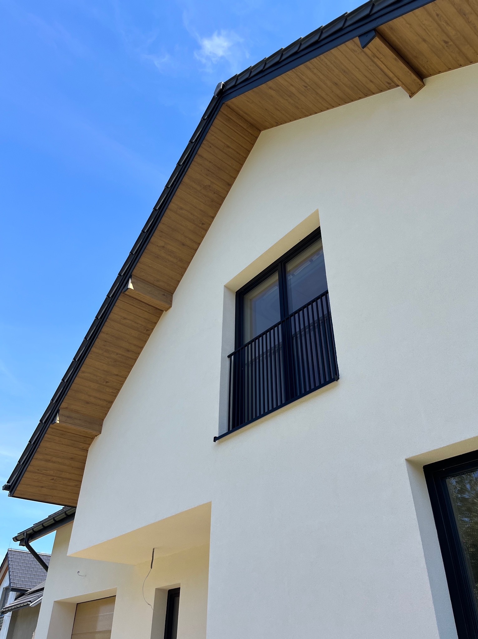 Biała elewacja domu jednorodzinnego z drewnianą podbitką i czarnymi ramami okiennymi, widziana pod kątem z dołu na tle błękitnego nieba.
