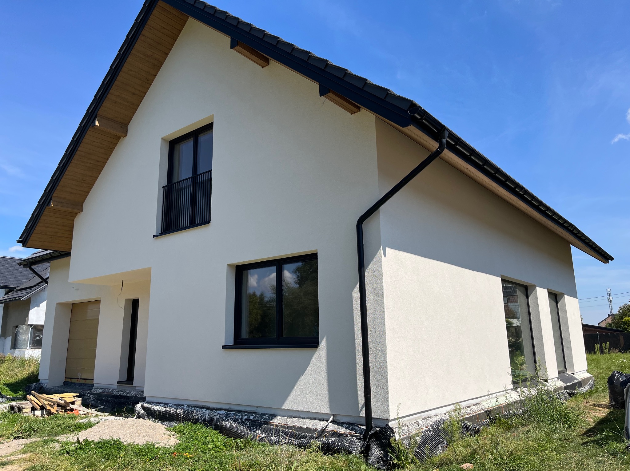 Nowoczesny dom jednorodzinny z jasną elewacją i czarnymi ramami okiennymi, widoczny z boku w słoneczny dzień. Uwagę zwraca wykończenie dachu z drewnianymi elementami.