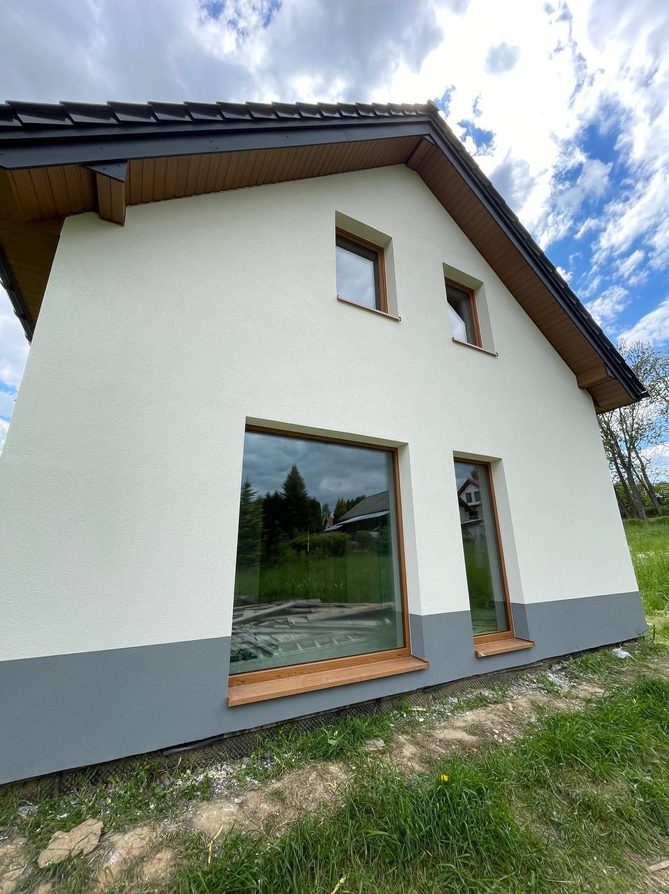 Biała elewacja domu jednorodzinnego z szarym cokołem, widok z dołu, okna z drewnianą stolarką, odbicie drzew w szybach, błękitne niebo z chmurami w tle.