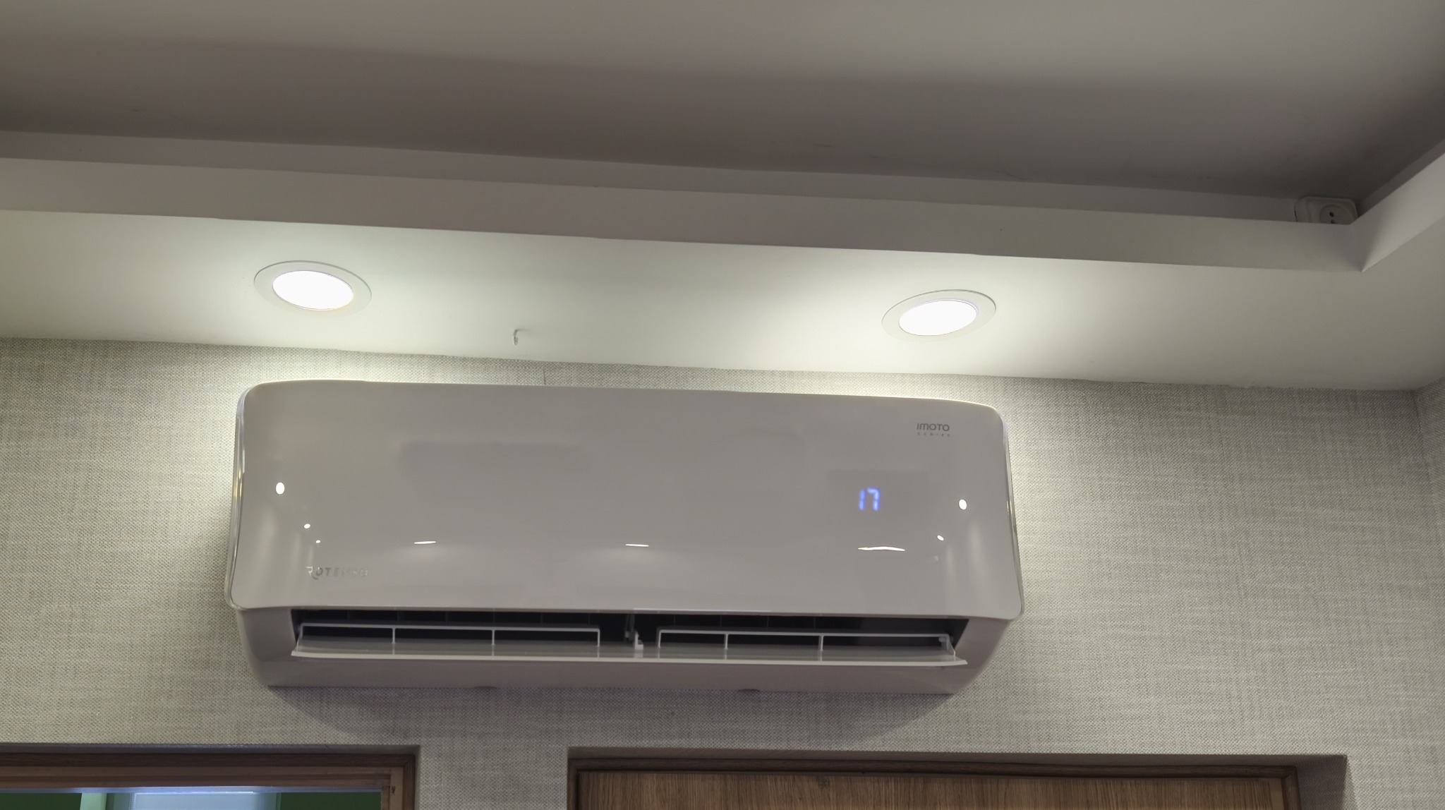 Biała klimatyzacja ścienna z wyświetlaczem temperatury 17 stopni Celsjusza, zamontowana na ścianie o szarej, teksturowanej tapecie w jasnym pomieszczeniu.