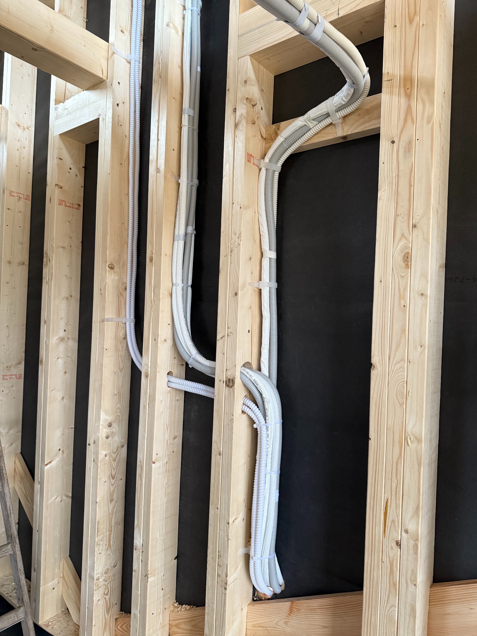 Białe, karbowane rury instalacyjne przymocowane opaskami do drewnianej konstrukcji szkieletowej domu na tle czarnej membrany, widoczne na etapie budowy.