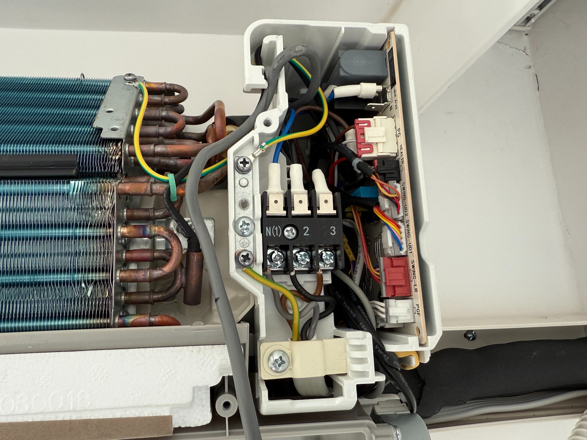 Otwarta obudowa klimatyzatora z widocznymi elementami wewnętrznymi: rurki miedziane, radiator z niebieskimi lamelami, panel sterowania z okablowaniem i złączkami.