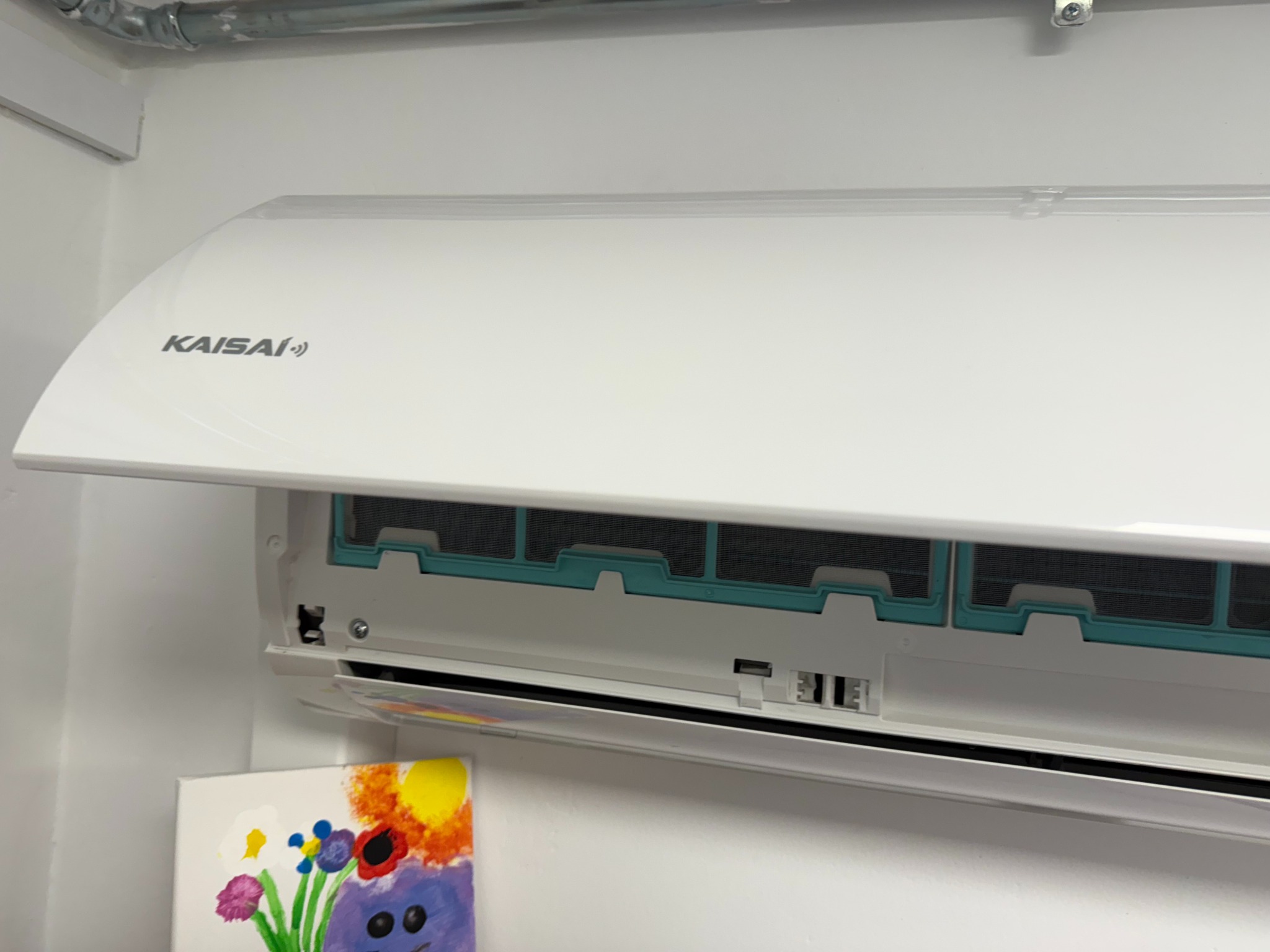 Ścienna jednostka klimatyzacyjna marki Kaisai z otwartą obudową, widoczne filtry, na ścianie obok obrazek z kwiatami.