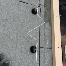KDF Construction Group Karalina Fabiianska - Montaż odgromu na płaskim dachu pokrytym papą, widoczne przyssawki i przewód odprowadzający, fragment buta roboczego w lewym dolnym rogu.