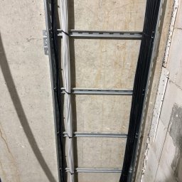 KDF Construction Group Karalina Fabiianska - Metalowa drabinka kablowa przymocowana do surowej betonowej ściany, wypełniona wiązkami białych i czarnych kabli elektrycznych, zabezpieczonych czarnymi opaskami zaciskowymi.
