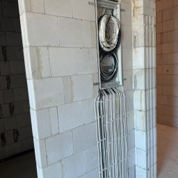 KDF Construction Group Karalina Fabiianska - Pionowy kanał kablowy z licznymi przewodami elektrycznymi prowadzonymi w ścianie z białych bloczków, widoczne zabezpieczenia kabli i napisy ołówkiem na ścianie.