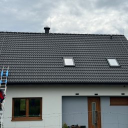 KDF Construction Group Karalina Fabiianska - Pracownik na drabinie przy nowo wybudowanym domu z ciemną dachówką i oknami dachowymi, widoczne zabezpieczenia odgromowe na dachu.