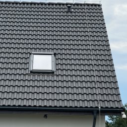KDF Construction Group Karalina Fabiianska - Fragment dachu z ciemną dachówką ceramiczną, okno dachowe, system odgromowy z metalowymi uchwytami oraz rynna w kolorze grafitowym.