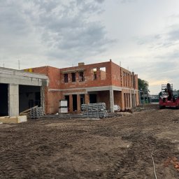 KDF Construction Group Karalina Fabiianska - Surowy stan budowy dwukondygnacyjnego domu z czerwonej cegły i betonu, widoczne rusztowania i materiały budowlane, ziemisty teren budowy z wyraźnymi śladami opon, pochmurne niebo w tle, widoczny...