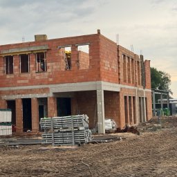 KDF Construction Group Karalina Fabiianska - Dwukondygnacyjny dom w trakcie budowy z czerwonej cegły, widoczne zbrojenia i szalunki, stosy rusztowań na pierwszym planie, ziemia z widocznymi śladami opon.