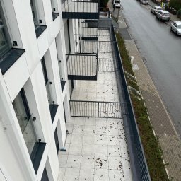 KDF Construction Group Karalina Fabiianska - Wysoki budynek z balkonami wyłożonymi jasnymi płytkami, widok z góry na balustrady, elewację z białych paneli i ulicę z zaparkowanymi samochodami, kilka opadłych liści na płytkach.