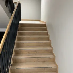 Schody wyłożone płytkami imitującymi drewno, z czarnymi paskami antypoślizgowymi i czarną metalową balustradą z drewnianym poręczą, na tle szarej ściany.