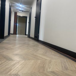 KDF Construction Group Karalina Fabiianska - Korytarz z jasną podłogą ułożoną w jodełkę, białe ściany z czarnymi listwami przypodłogowymi, czarne drzwi i oświetlenie sufitowe w perspektywie zbieżnej.