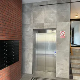 Nowoczesny hol wejściowy z windą, wykończony płytami imitującymi marmur i cegłą, z czarnymi skrzynkami pocztowymi i wzorzystą posadzką.