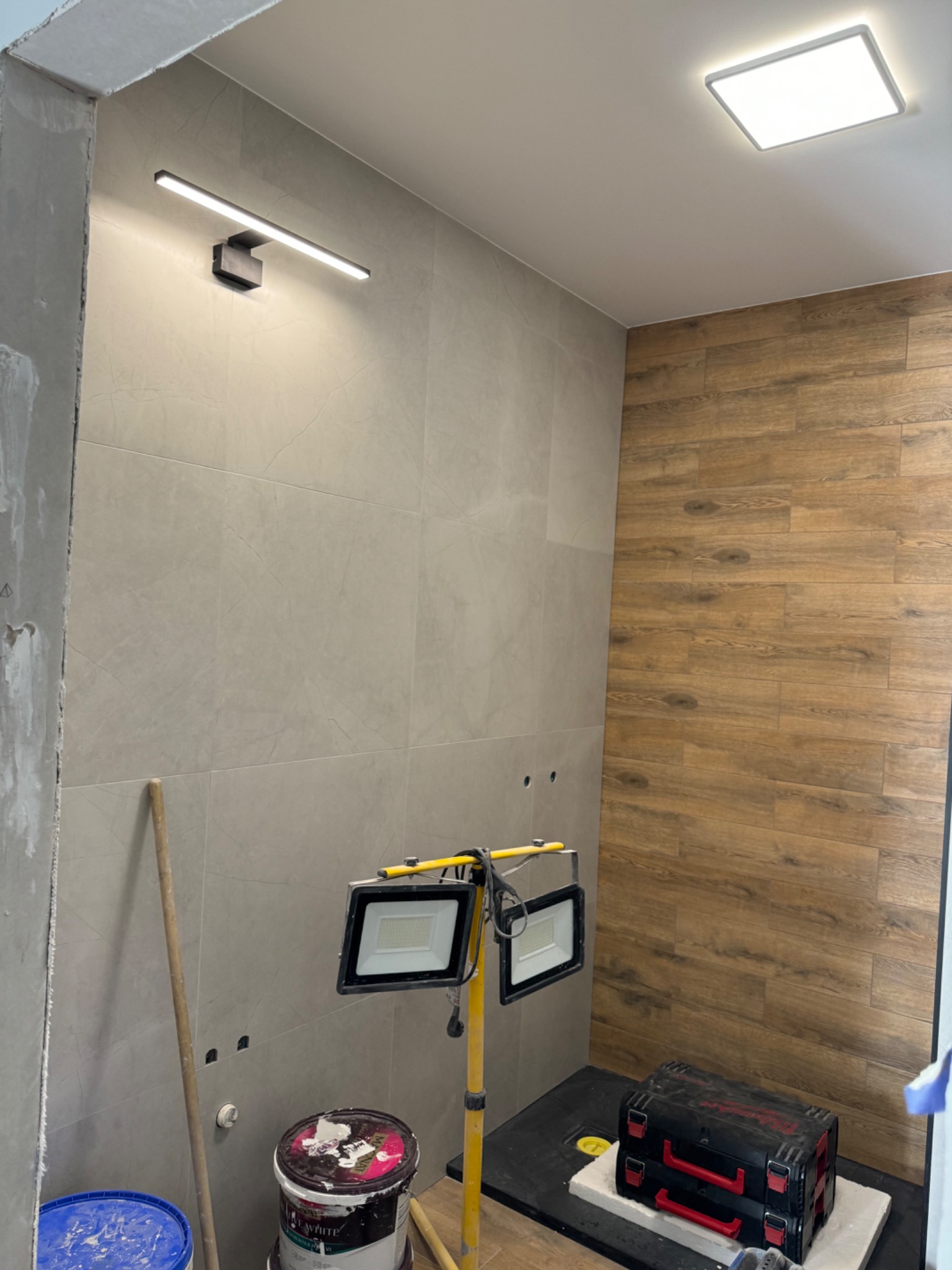 Łazienka w trakcie remontu: szare płytki ścienne z delikatnym wzorem, drewnopodobne płytki na ścianie, lampa LED nad lustrem, reflektory halogenowe na statywie, wiadro z farbą i skrzynka narzędziowa.