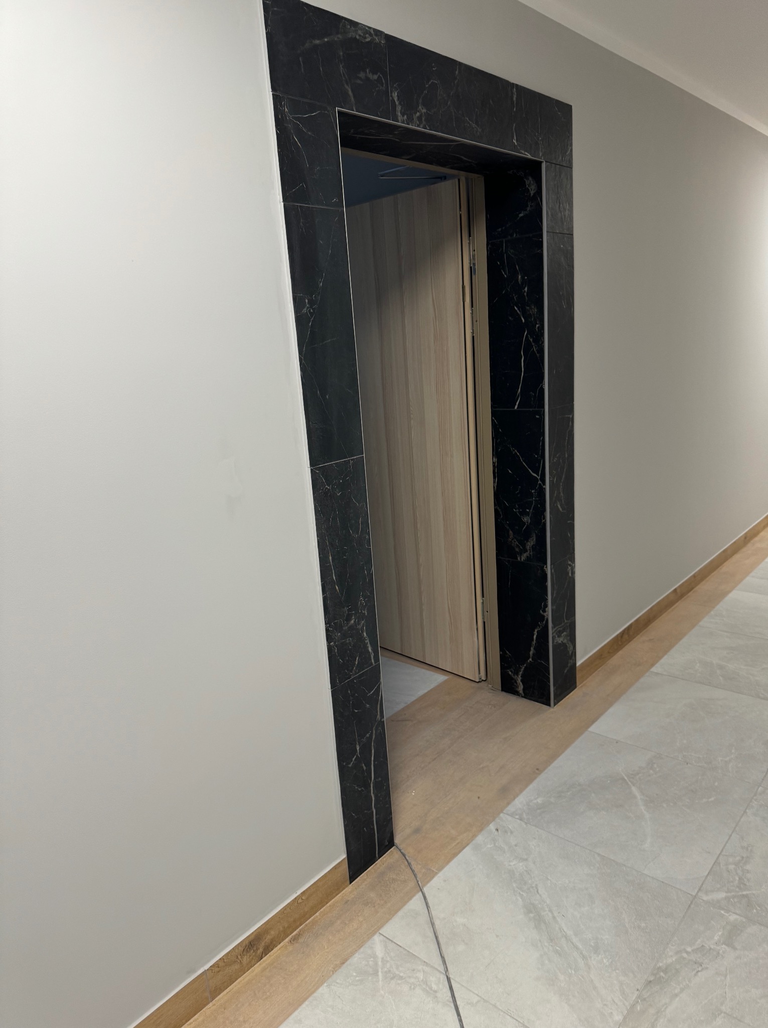 Wykończone wejście do pokoju z ościeżnicą z czarnego marmuru, szara ściana i podłoga w odcieniach szarości i beżu.