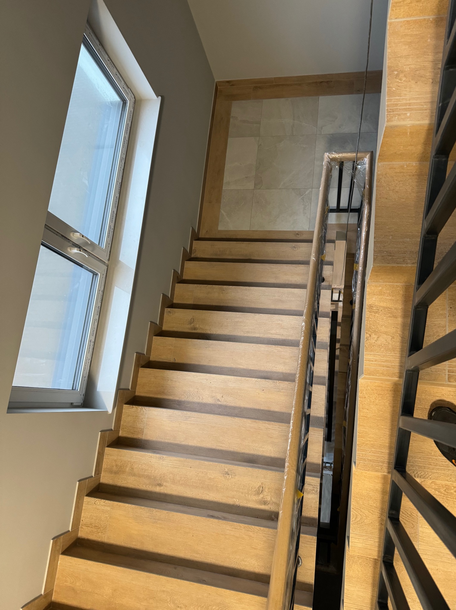 Schody drewniane z metalową balustradą, widok z góry, nowoczesne wykończenie, duże okno doświetlające przestrzeń, balustrada zabezpieczona folią ochronną.