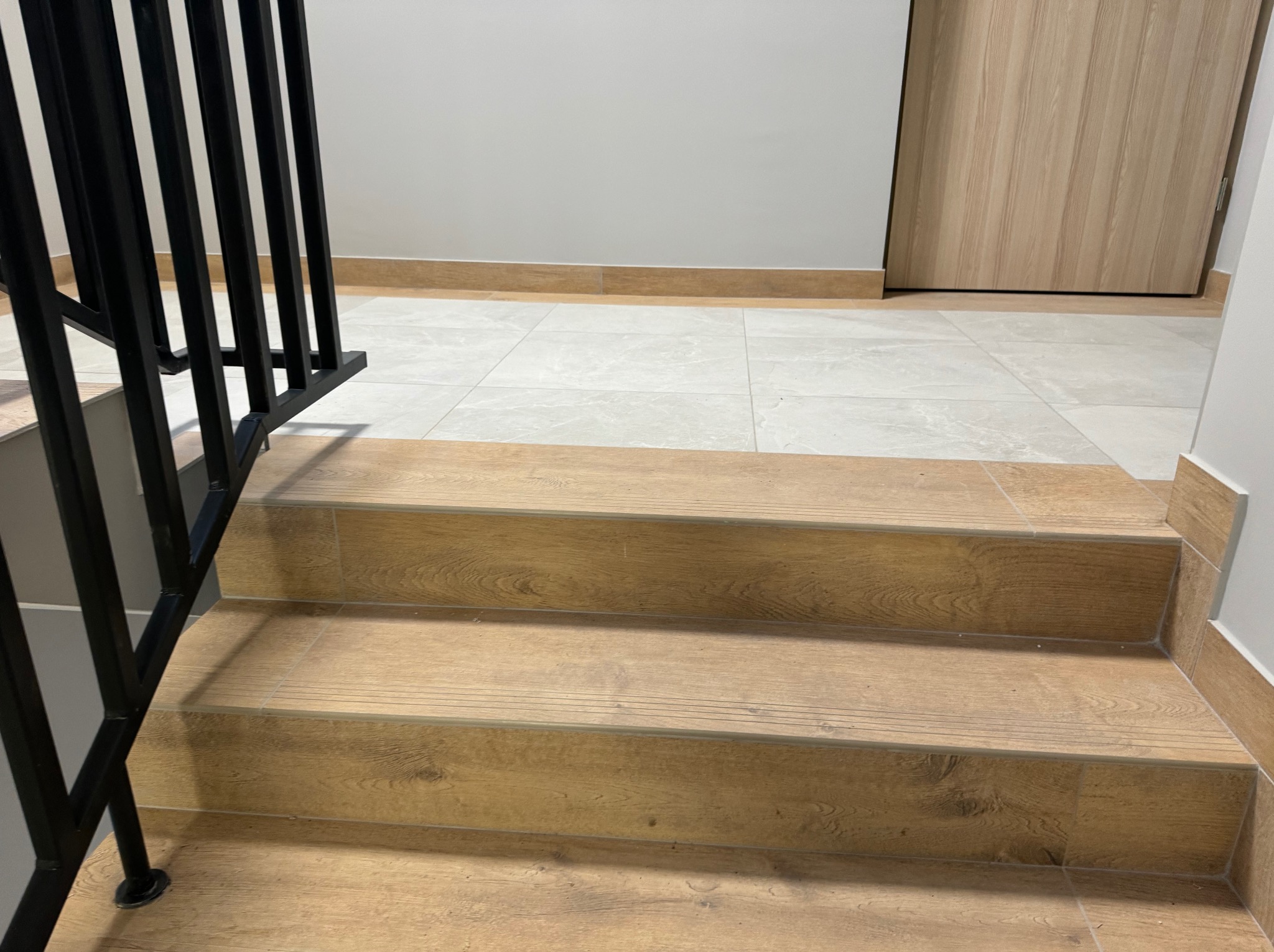 Klatka schodowa wyłożona płytkami imitującymi drewno, z czarną metalową balustradą, prowadząca do drzwi w tle. Płytki podłogowe w odcieniach szarości. Widok z dołu schodów.