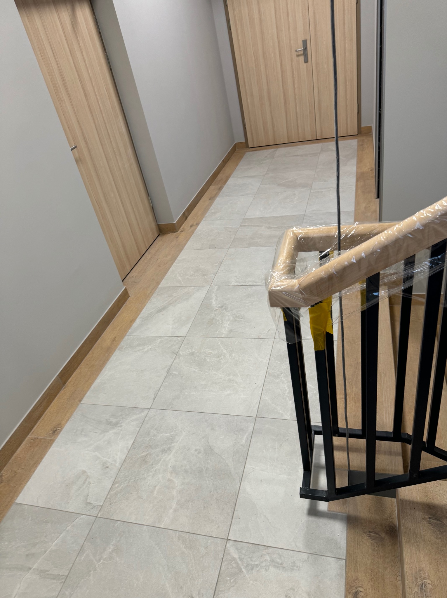 Korytarz z jasnymi, kwadratowymi płytkami podłogowymi, drewnianymi drzwiami i czarną, metalową balustradą schodów z drewnianym poręczą owiniętym folią ochronną.