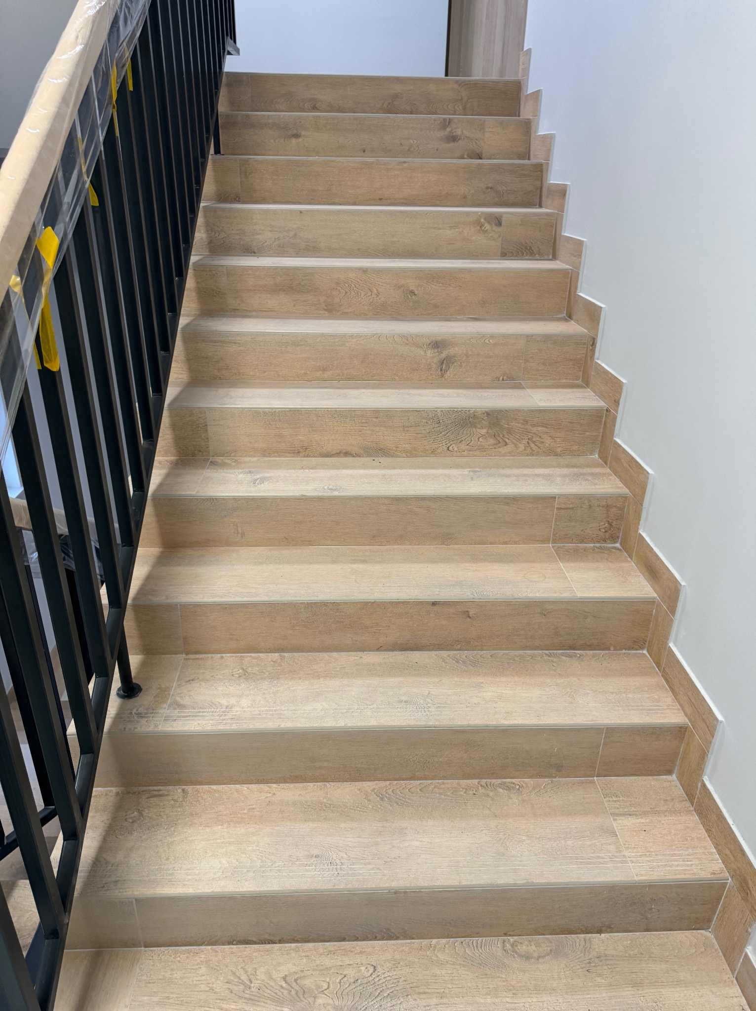 Schody wewnętrzne wyłożone płytkami imitującymi drewno, z czarną, metalową balustradą, zabezpieczoną folią ochronną na górnej części poręczy.