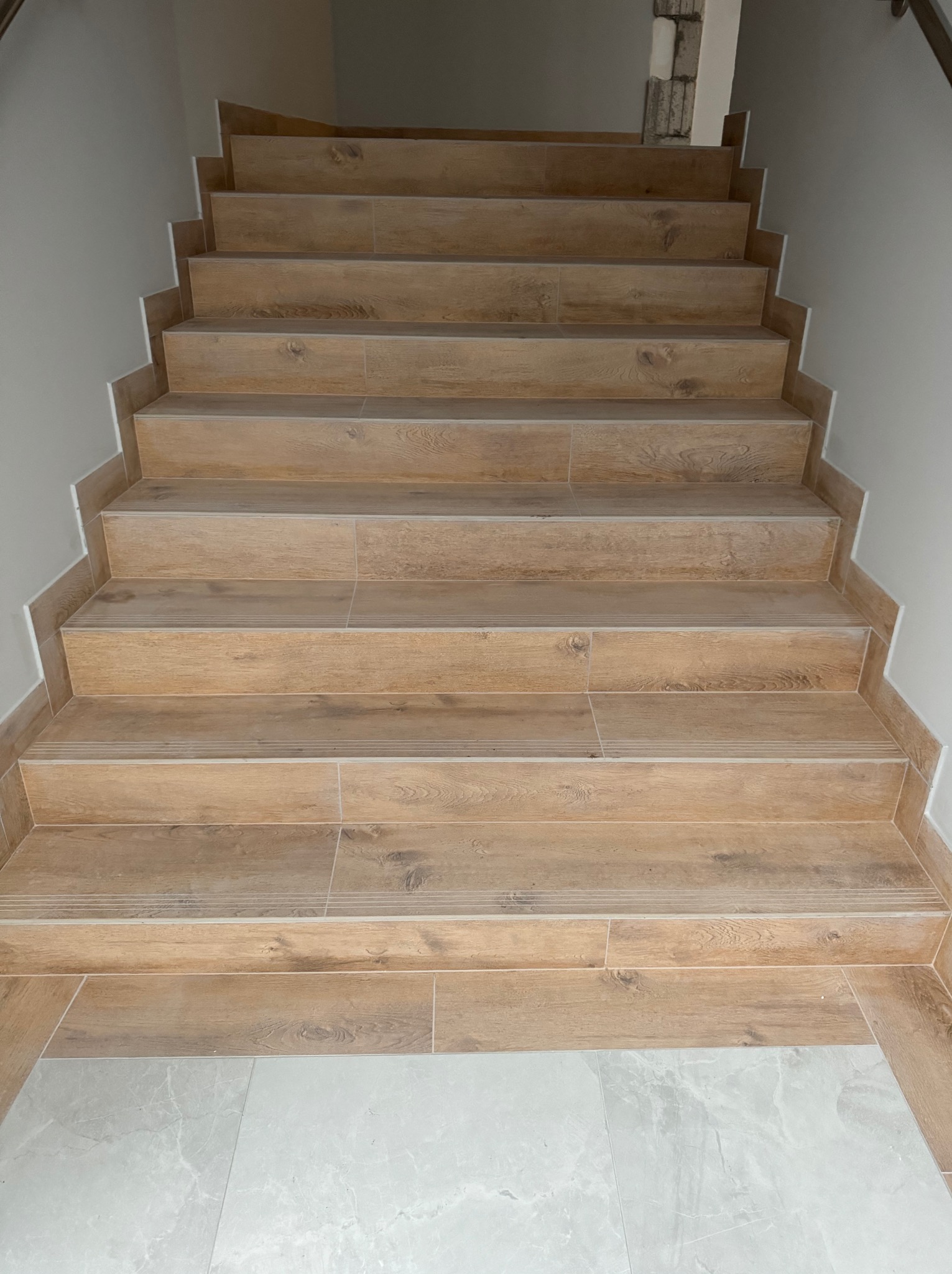 Schody wykończone płytkami imitującymi drewno, z delikatnymi białymi fugami i listwami przypodłogowymi, prowadzące w górę; na dole widoczne szare płytki podłogowe.