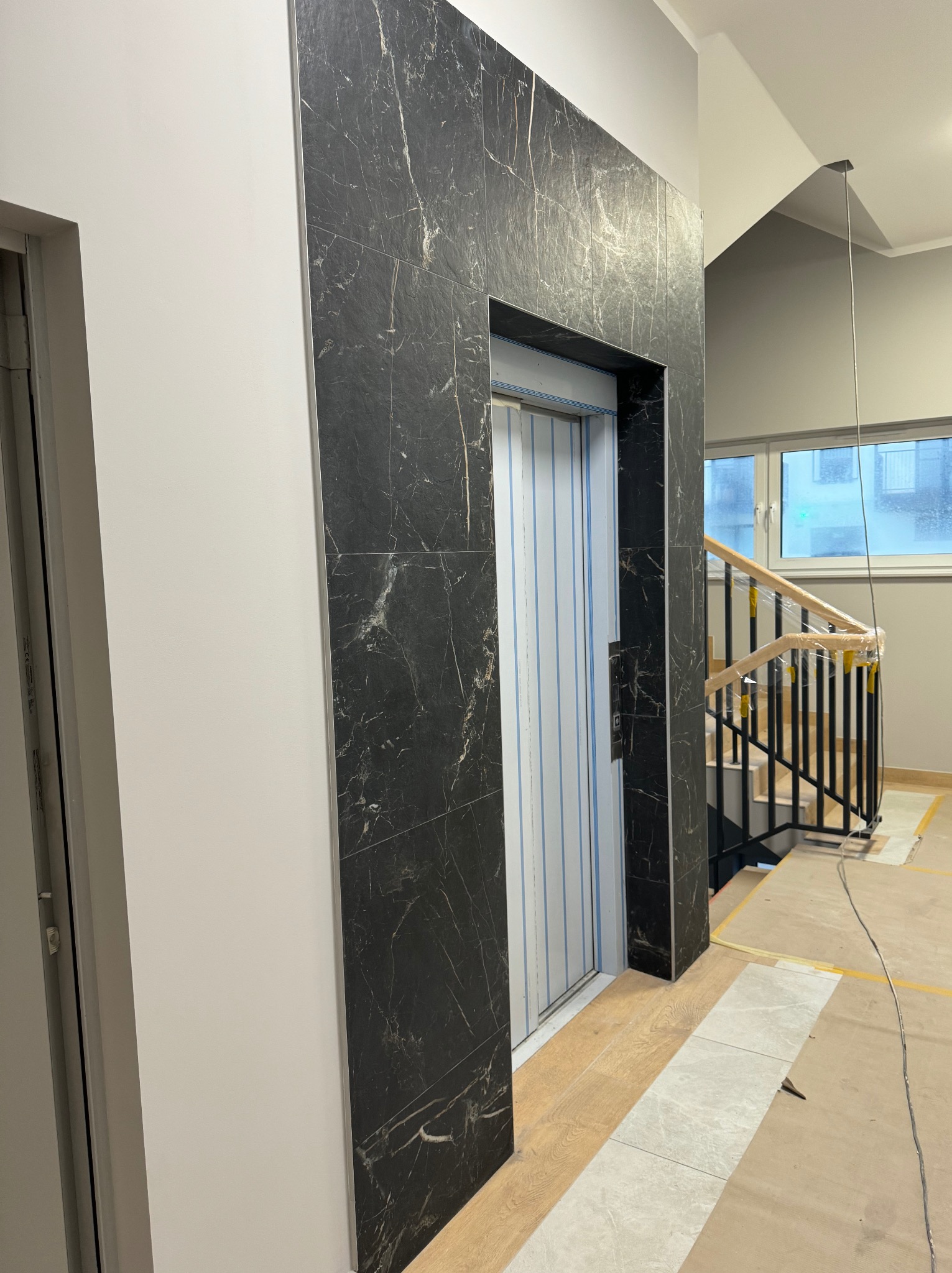 Wnętrze klatki schodowej w trakcie wykańczania z widoczną windą obłożoną czarnym marmurem, schodami z czarną balustradą i drewnianymi poręczami zabezpieczonymi folią oraz częściowo położoną podłogą.